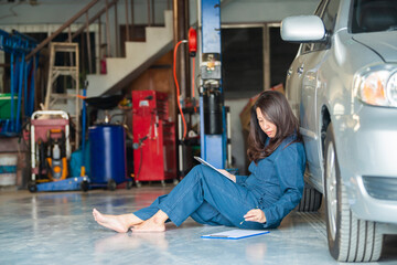 Wall Mural - Asian woman mechanic using tablet to fix car and writing to a clipboard while sitting on the floor at the repair garage. Copy space