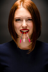 Happy eyelash master holding eyelash extension eyebrush in her teeth, looking at camera on dark backdrop.