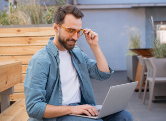Young successful programmer using laptop computer, working outdoors. Smiling student studying, distance learning, online education concept  