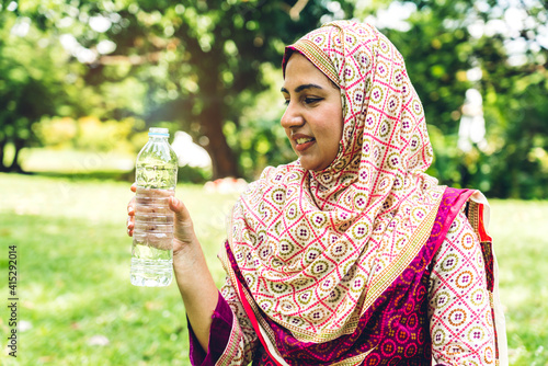 Portrait of happy arabic muslim woman with hijab dress drinking water from a bottle while relaxing and feeling fresh on green natural background at summer green park.Healthy lifestyle concept