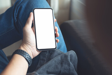 Wall Mural - Mockup image of mobile smartphone with blank screen template, man sitting on sofa, hand holding and looking at mobile phone screen