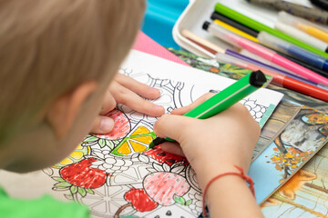 Over the shoulder: a boy learns to color a coloring book with fruits and vegetables with a green felt-tip pen. Development of creative thinking at home.