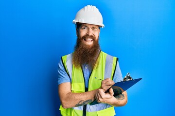 Canvas Print - Redhead man with long beard wearing safety helmet holding clipboard winking looking at the camera with sexy expression, cheerful and happy face.