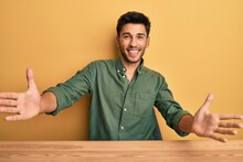 Young Handsome Man Wearing Casual Clothes Sitting On The Table Looking At The Camera Smiling With Open Arms For Hug. Cheerful Expression Embracing Happiness.