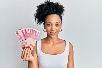 Poster - Young african american girl holding indonesian rupiah banknotes looking positive and happy standing and smiling with a confident smile showing teeth