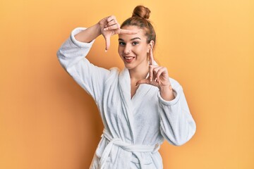 Wall Mural - Young blonde woman wearing bathrobe smiling making frame with hands and fingers with happy face. creativity and photography concept.