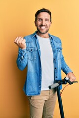 Wall Mural - Young handsome man standing on modern scooter over yellow background screaming proud, celebrating victory and success very excited with raised arm