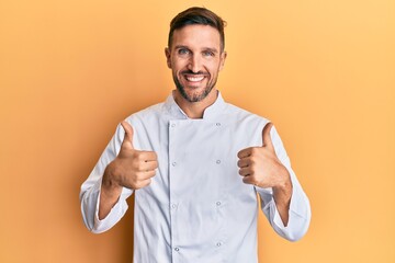 Canvas Print - Handsome man with beard wearing professional cook uniform success sign doing positive gesture with hand, thumbs up smiling and happy. cheerful expression and winner gesture.