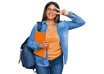 Poster - Young latin girl wearing student backpack and holding books doing peace symbol with fingers over face, smiling cheerful showing victory