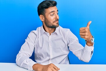 Handsome hispanic man wearing business clothes sitting on the table looking proud, smiling doing thumbs up gesture to the side