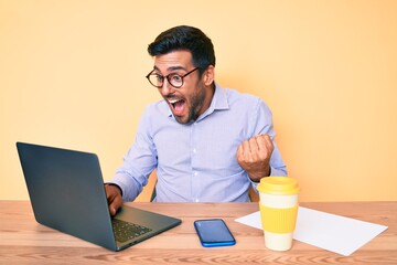 Canvas Print - Young hispanic man working at the office drinking a cup of coffee screaming proud, celebrating victory and success very excited with raised arms