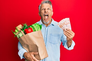 Wall Mural - Middle age grey-haired man holding groceries and singapore dollars banknotes angry and mad screaming frustrated and furious, shouting with anger looking up.