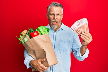 Sticker - Middle age grey-haired man holding groceries and new zealand dollars banknotes in shock face, looking skeptical and sarcastic, surprised with open mouth