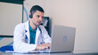 © alex_marina - Friendly Male Doctor in White Medical Coat With Headphones Making Conference Call on Laptop.Remote Consulting Patient Online From Healthcare Hospital. Telemedicine.