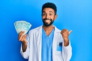 Poster - Handsome hispanic man with beard wearing doctor uniform holding 100 brazilian reals pointing thumb up to the side smiling happy with open mouth