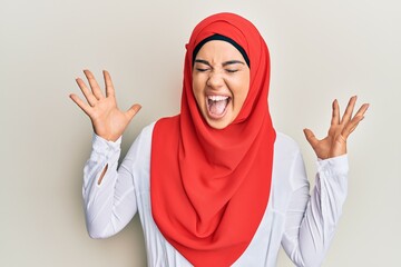 Wall Mural - Young beautiful hispanic girl wearing traditional islamic hijab scarf celebrating mad and crazy for success with arms raised and closed eyes screaming excited. winner concept