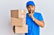© Krakenimages.com - Handsome man with beard wearing courier uniform holding delivery packages asking to be quiet with finger on lips. silence and secret concept.
