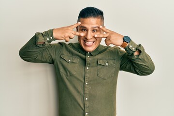 Young latin man wearing casual clothes doing peace symbol with fingers over face, smiling cheerful showing victory