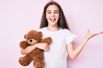 Cute hispanic child girl holding teddy bear celebrating victory with happy smile and winner expression with raised hands