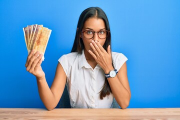 Canvas Print - Beautiful hispanic woman holding 500 norwegian krone banknotes covering mouth with hand, shocked and afraid for mistake. surprised expression