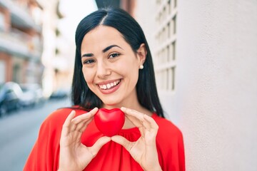 Wall Mural - Young latin girl smiling happy holding heart at the city.