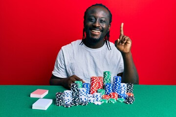 Sticker - Handsome young black man sitting on the table with poker chips and cards pointing finger up with successful idea. exited and happy. number one.