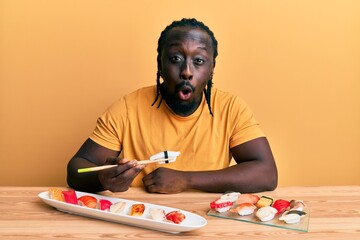 Poster - Handsome young black man eating sushi sitting on the table scared and amazed with open mouth for surprise, disbelief face