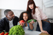 © sofiko14 - Blurred view of happy mixed race teen girl daughter sitting between parents and showing fresh vegetables tomato and cucumber to camera. Focus on vegetables