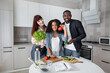 © sofiko14 - Cheerful young pleasant multiethnic family with cute daughter, showing fresh vegetables, cucumber, tomato and lettuse to the camera, posing at home kitchen