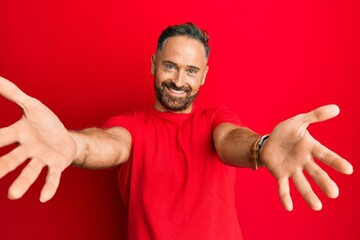 Handsome middle age man wearing casual red tshirt looking at the camera smiling with open arms for hug. cheerful expression embracing happiness.