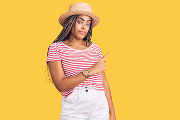 Canvas Print - Young african american woman with braids wearing summer hat pointing with hand finger to the side showing advertisement, serious and calm face