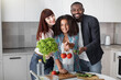 © sofiko14 - Multiracial family showing vegetables to the camera, posing in light modern kitchen interior at home. Family cooking, healthy food concept