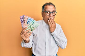 Poster - Middle age indian man holding thai baht banknotes covering mouth with hand, shocked and afraid for mistake. surprised expression