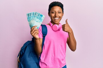 Wall Mural - Young african american girl wearing student backpack and holding brazilian real banknotes smiling happy and positive, thumb up doing excellent and approval sign