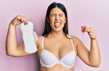 Poster - Beautiful brunette woman holding sanitary pad and reusable menstruation cup sticking tongue out happy with funny expression.