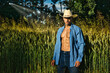 Portrait of sexy farmer or cowboy in hat with unbuttoned shirt on muscular torso, looking at camera, while standing next to hay field in countryside