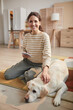 © Seventyfour - Vertical full length portrait of smiling young woman looking at camera while sitting on floor with pet dog in cozy home interior