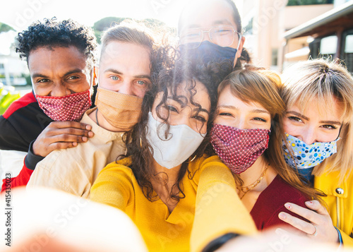 Multicultural friends covered by face masks taking a selfie outdoor - New normal friendship concept with young people smiling and having fun - Bright filter