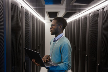 Side view portrait of young African American data engineer working with supercomputer in server room and holding laptop, copy space