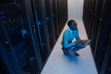 Wall Mural - High angle view at young African American data engineer working with supercomputer in server room lit by blue light, copy space