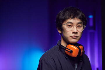 Wall Mural - Portrait of young Asian man wearing headphones looking at camera while standing in futuristic interior lit by blue light, copy space