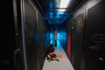Wall Mural - Wide angle portrait of young Asian man using laptop while sitting on floor in server room lit by blue light and setting up supercomputer network , copy space