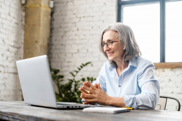 Senior businesswoman sitting at the desk, looking at webcam and talking with client online or watching tutorial, elder female office worker or therapist having appointment, virtual meeting via laptop