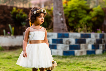 Portrait of an Asian, Indian female child dressed as a flower girl in a white dress and floral hair band and holding a bouquet outdoors in a flower park