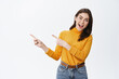 © Cookie Studio - Excited attractive caucasian woman pointing fingers left, showing advertisement and inviting to check it out. Girl with joyful face demonstrating promo, white background