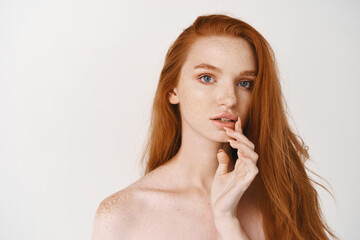 Wall Mural - Close-up of gorgeous redhead lady with pale smooth skin and no makeup, touching lips and looking at camera thoughtful, standing naked on white background