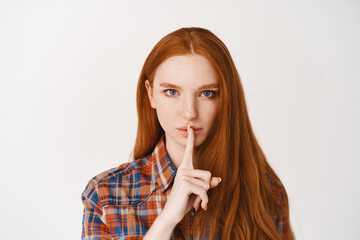 Wall Mural - Serious young lady with red natural hair, shushing at camera, telling a secret with confident face, standing over white background