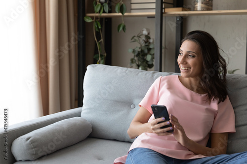 Young arab woman sit on comfy couch look at window with smile distracted from cell screen get pleasant message. Positive indian lady spend time online enjoy web shopping via phone dreaming. Copy space © fizkes