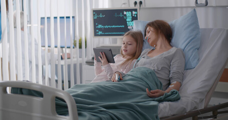 Little girl visiting sick mother in hospital showing photo on digital tablet