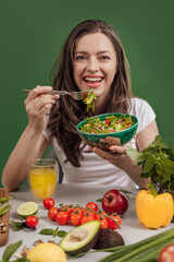 Portrait of a young cheerful woman at the table full of healthy raw vegetables and fruits on green background. Concept of vegetarianism, healthy eating and wellness
